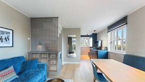 A modern living room with a blue sofa, a wooden dining table, and a kitchen area with a window. - Dependance Windrose 174 (Wenningstedt-Braderup (Sylt))