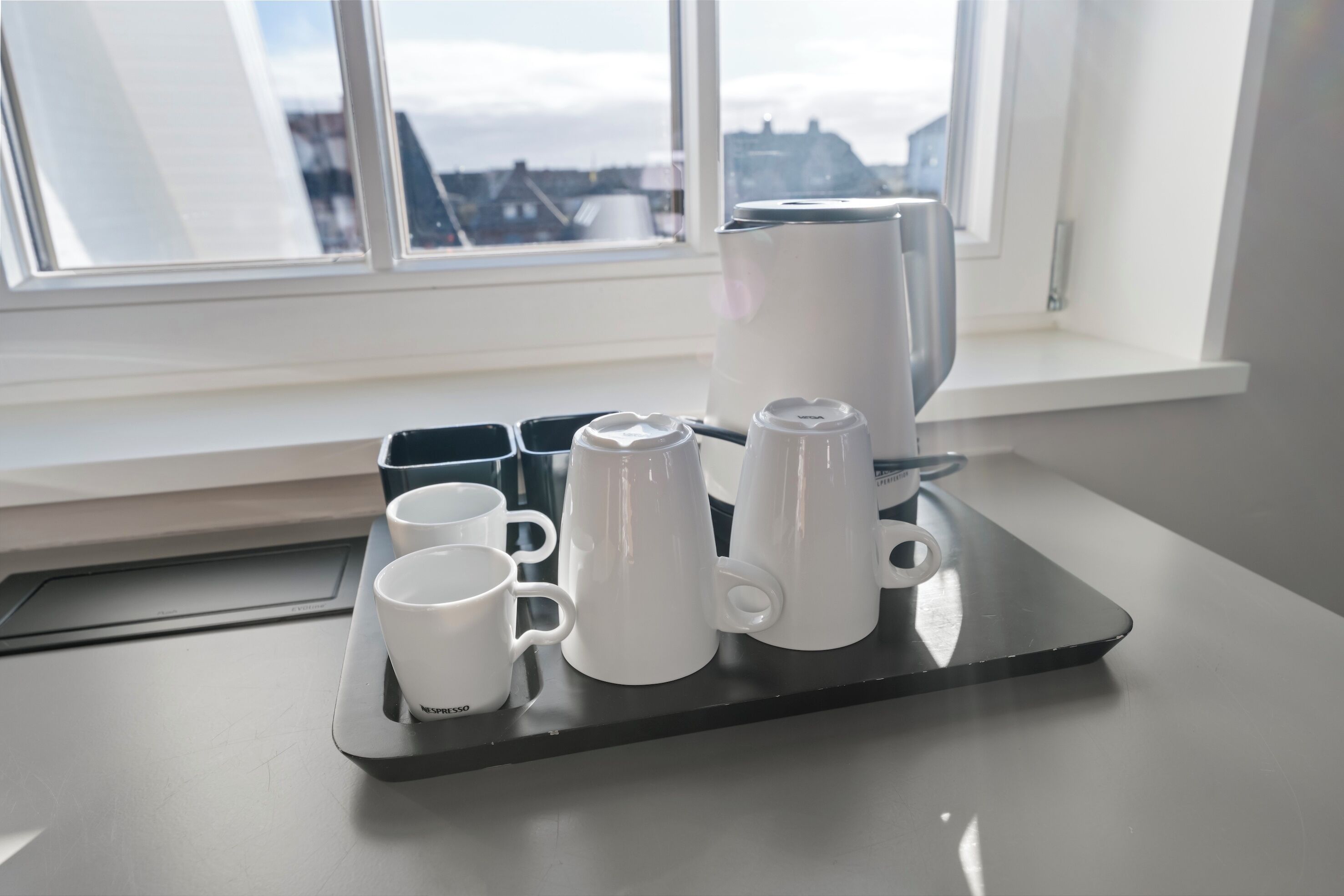 Una bandeja con tazas blancas y una cafetera sobre una mesa junto a una ventana.