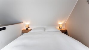 Ein Schlafzimmer mit schrägverlaufender Decke, einem Bett mit weißer Bettwäsche, zwei Nachttischen mit Lampen und einem kleinen Nachttisch aus Holz.