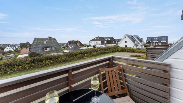 Ein Balkon mit Tisch und zwei Gläsern Weißwein, mit Blick auf ein Wohngebiet mit Häusern und blauem Himmel.