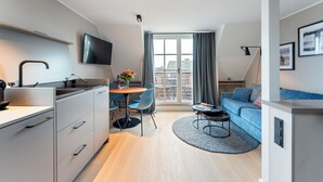 Eine moderne Wohnung mit Küche, Essbereich und Wohnzimmer. Ein großes Fenster mit Vorhängen, ein flachbildfernseher an der Wand und eine Couch mit Kissen.