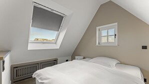A bedroom with a sloped ceiling, a skylight, a small window, a radiator, and a bed with white bedding. - Dependance Windrose 274 (Wenningstedt-Braderup (Sylt))