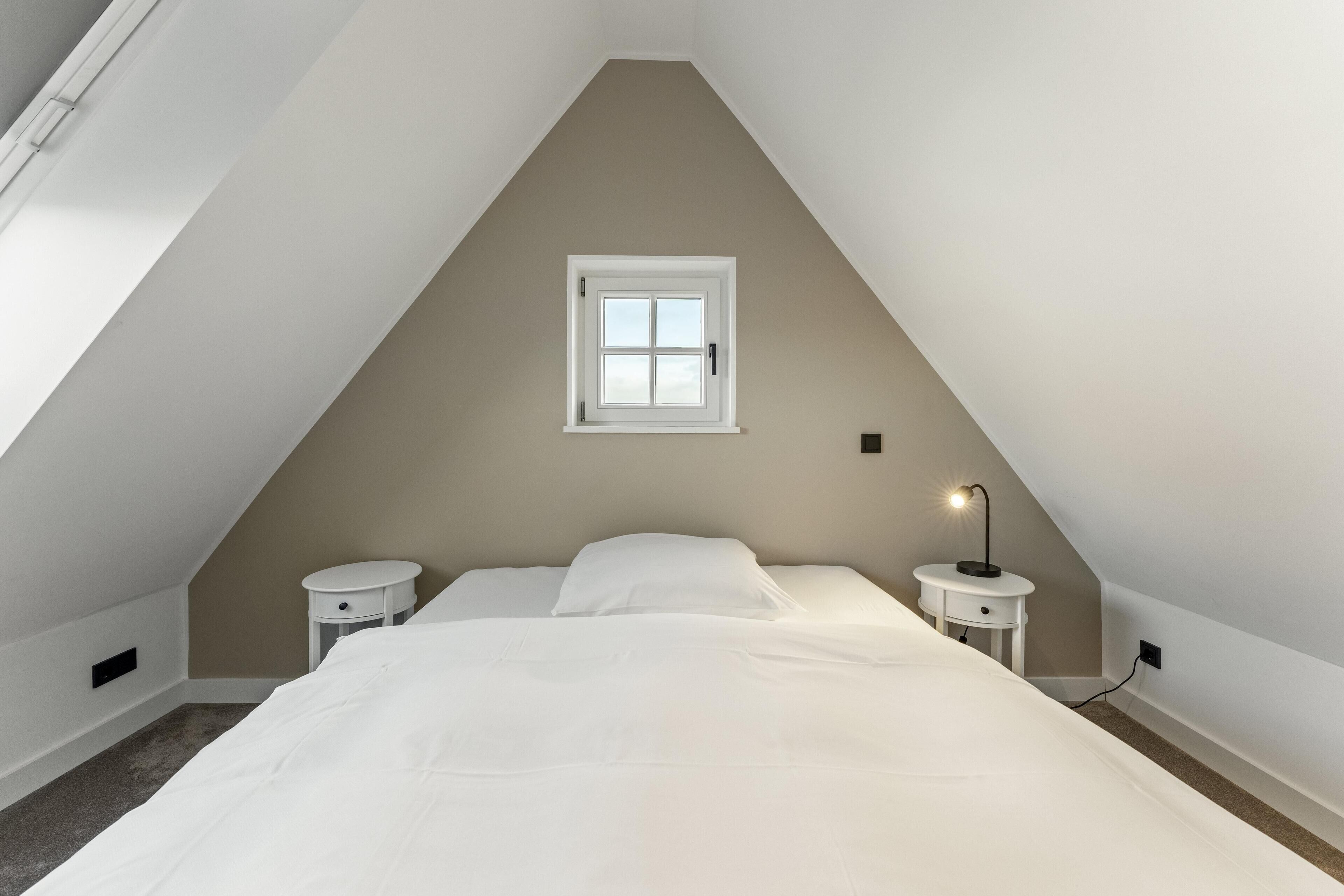 A bedroom with a sloped ceiling, a single window, two bedside tables, a bed with white bedding, and a nightstand with a lamp.