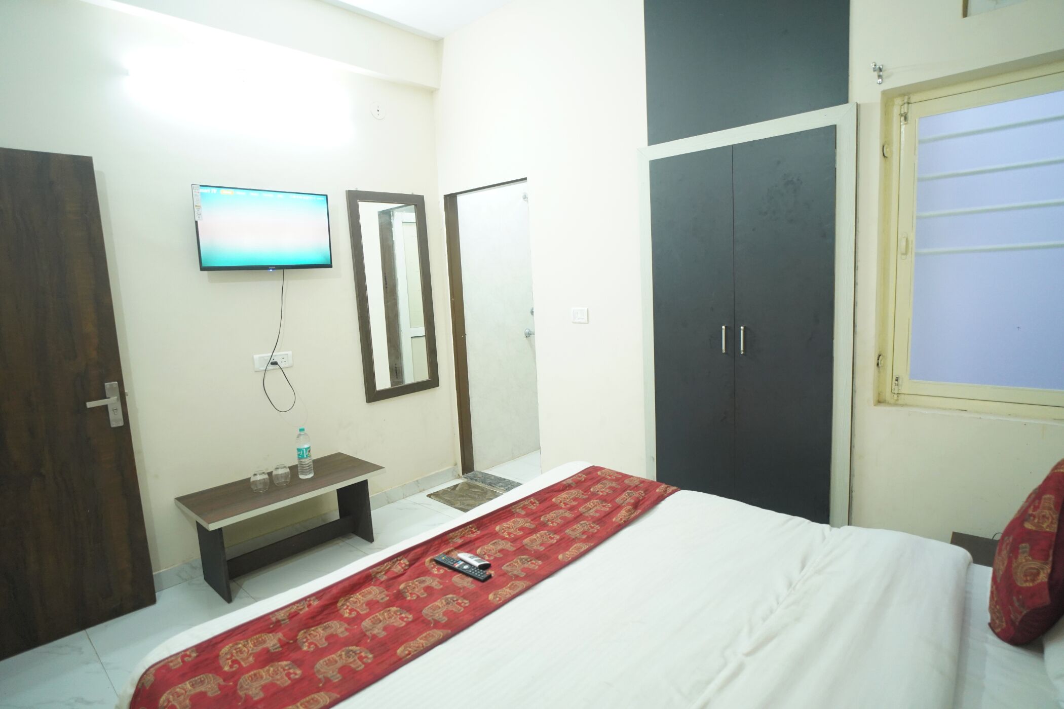 A hotel room with a bed, a TV mounted on the wall, a bench, and a wardrobe.