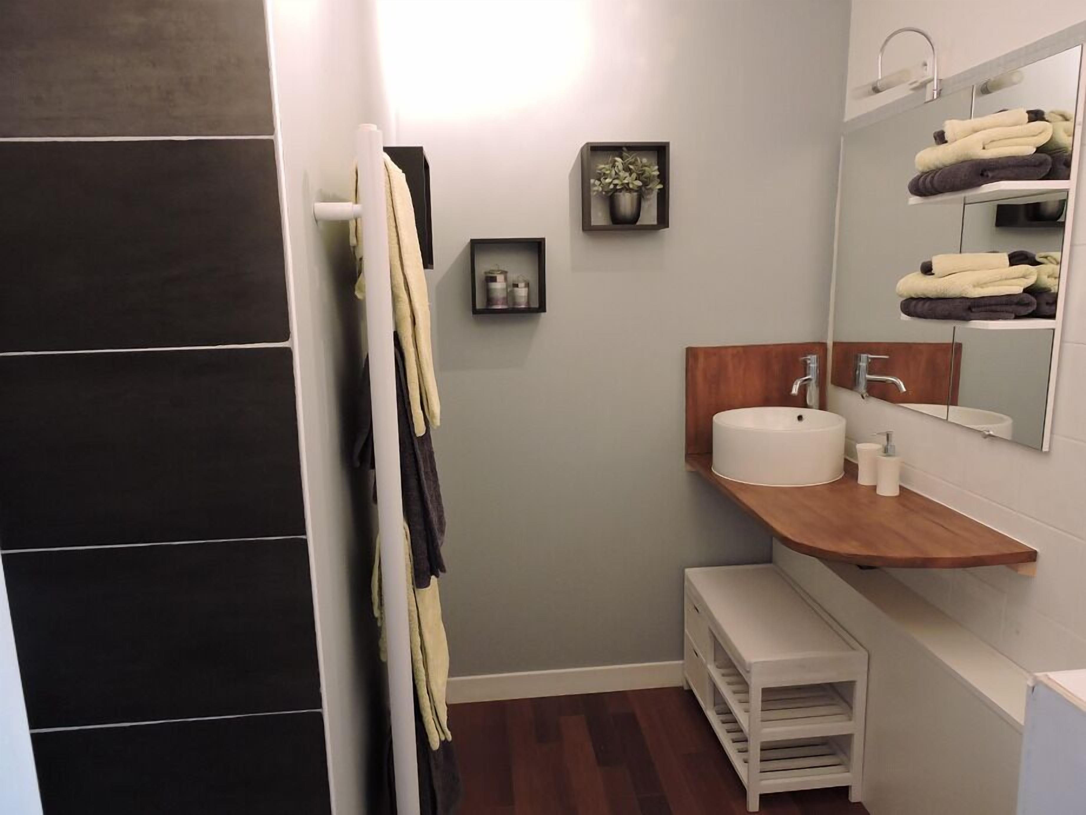 A modern bathroom with a dark tiled wall, a wooden vanity, a sink, a mirror, and a towel rack.