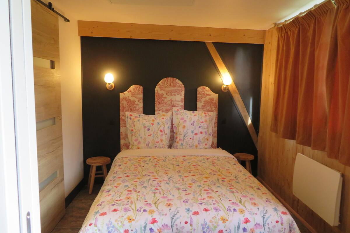 Un dormitorio con colcha estampada, cabecera de madera, dos mesitas de noche y una pared oscura con dos luces empotradas.
