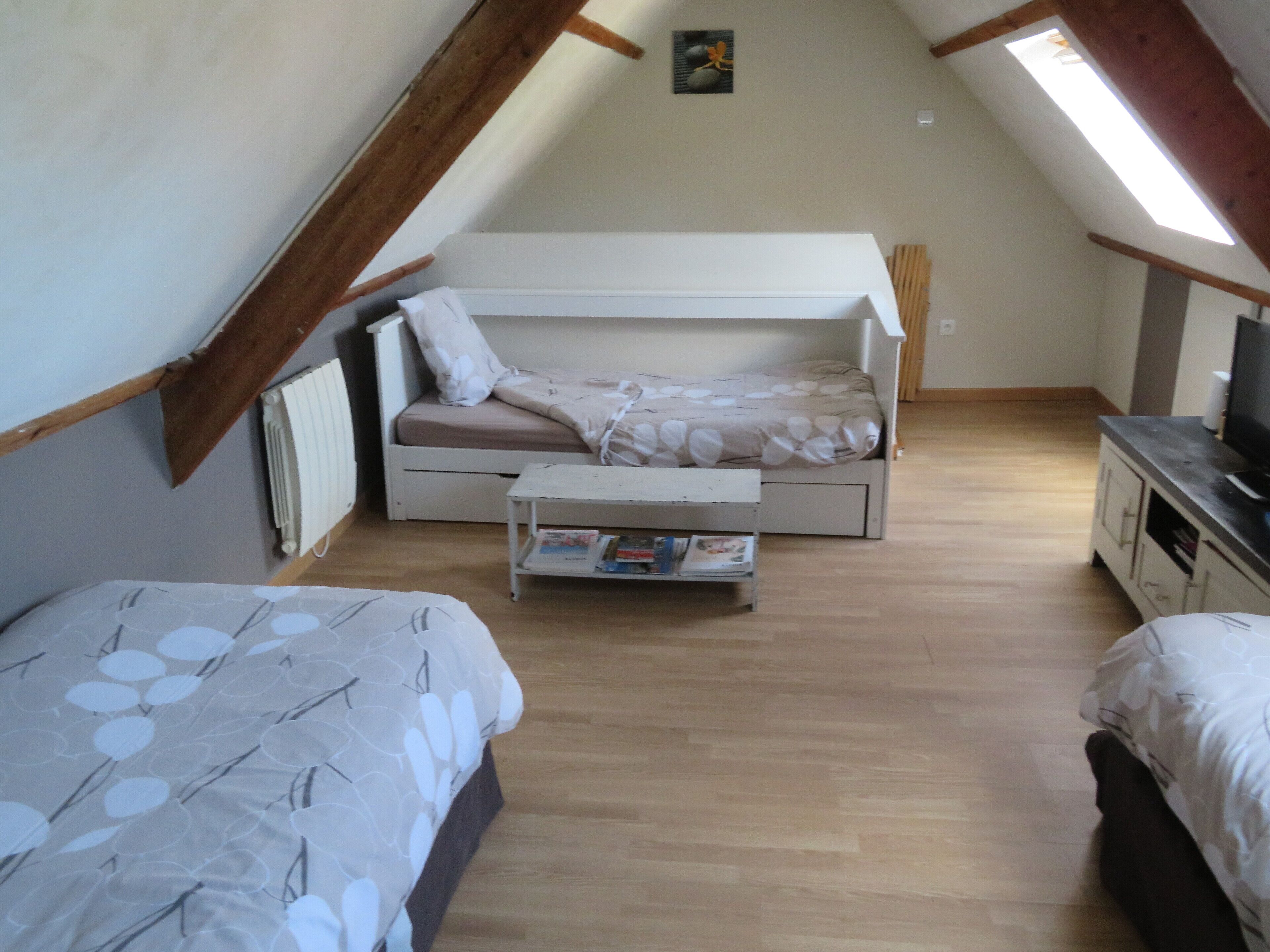 A small, cozy attic bedroom with a bunk bed, a single bed, a small table with books, and a television on a dresser.