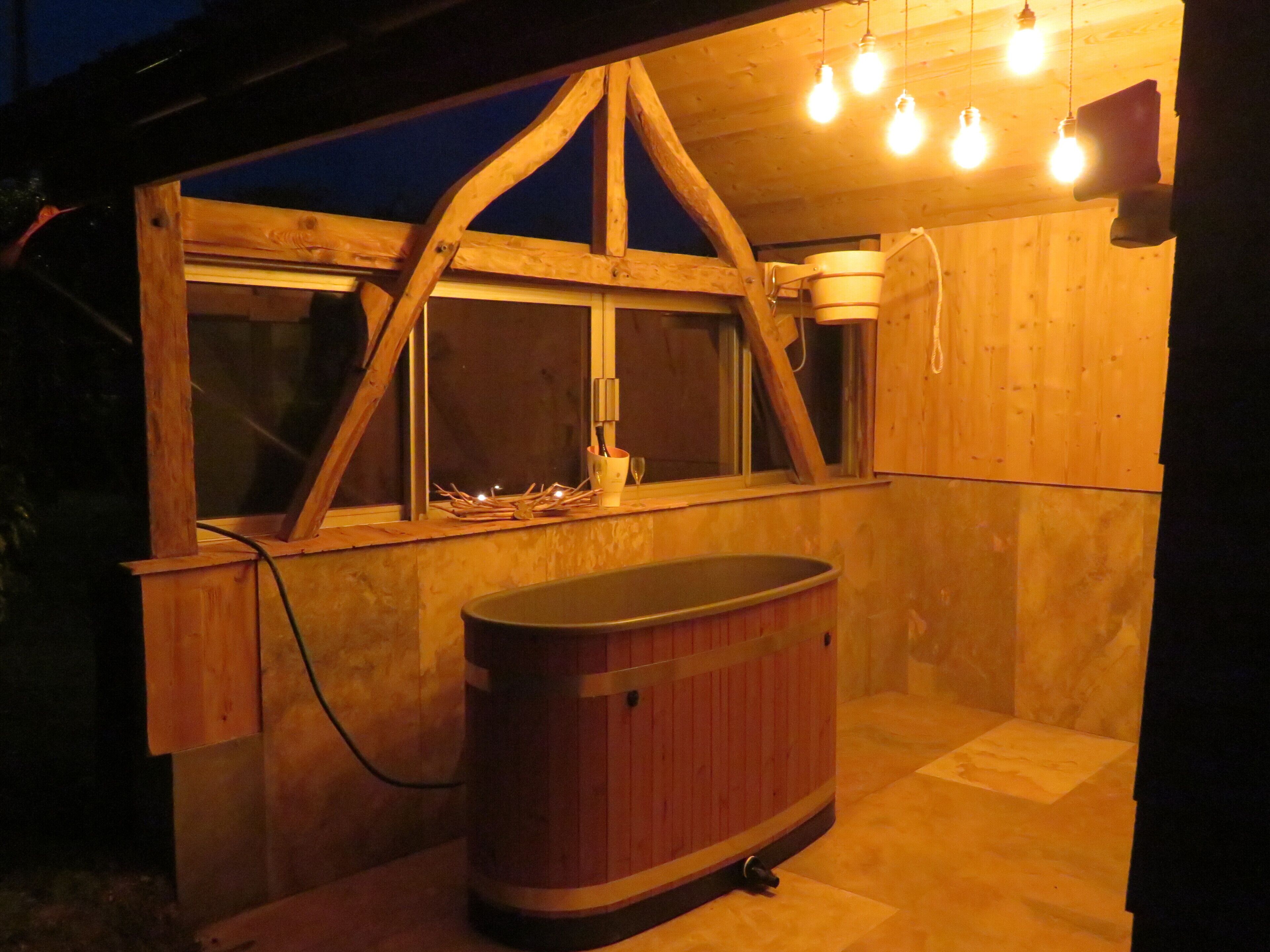 A wooden structure with a window, a hot tub, and hanging light bulbs.