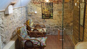 A cozy attic room with stone walls, wooden ceiling beams, and a sloped roof. There are wicker chairs with colorful cushions, a wooden bench, and a glass shower enclosure. - La linoterie Sauna privatif (Colembert)