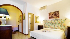 A bedroom with a bed, a nightstand, a dresser, a lamp, a door, and a painting on the wall. - Villa Miramì (Saturnia)