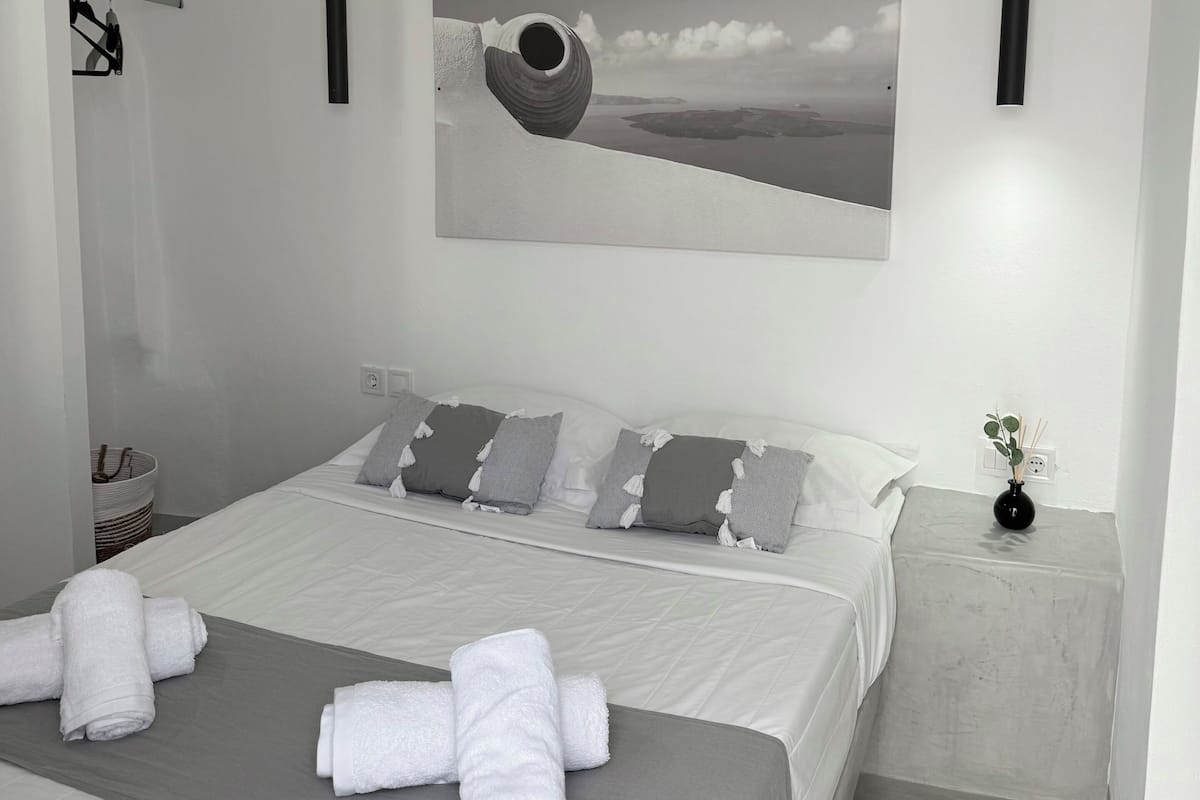 Een moderne slaapkamer met een groot bed, grijs beddengoed, een grijs hoofdbord, een grijze nachtkastje met een vaas, en een kunstwerk aan de muur.