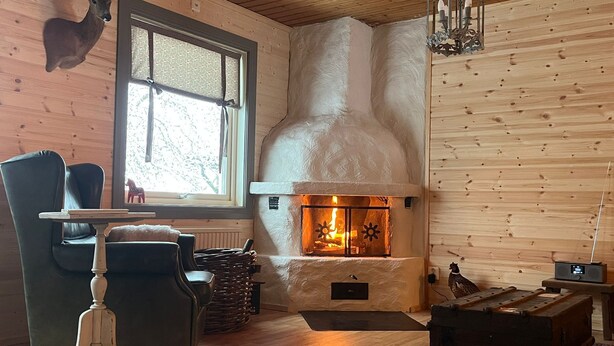 A cozy room with a fireplace, a wooden wall, a window, a chair, and a table. - Attractive guesthouse with spacious garden and extra service in the middle of nature (Lysvik)