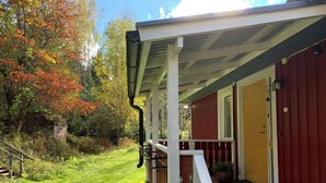 A wooden deck with a white railing, a red building with a yellow door, and a small shed in a wooded area. - Attractive guesthouse with spacious garden and extra service in the middle of nature (Lysvik)