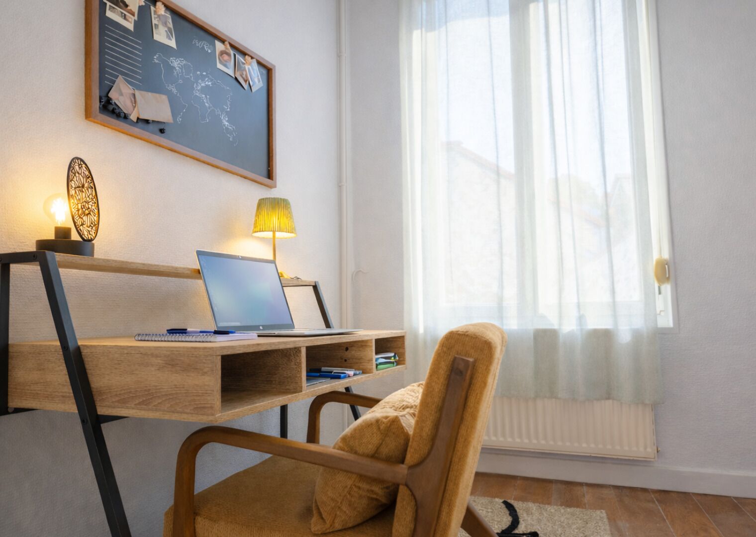 Werkplek met een laptop, bureau, stoel, lamp en prikbord met foto’s en aantekeningen.