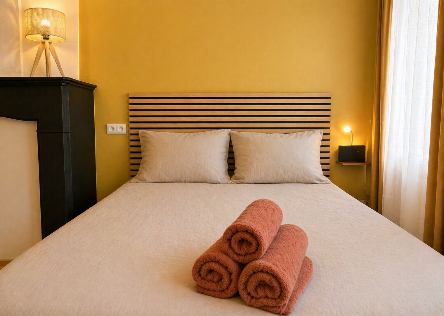 A hotel room with a bed, two pillows, a lamp, and a rolled towel on the bed.