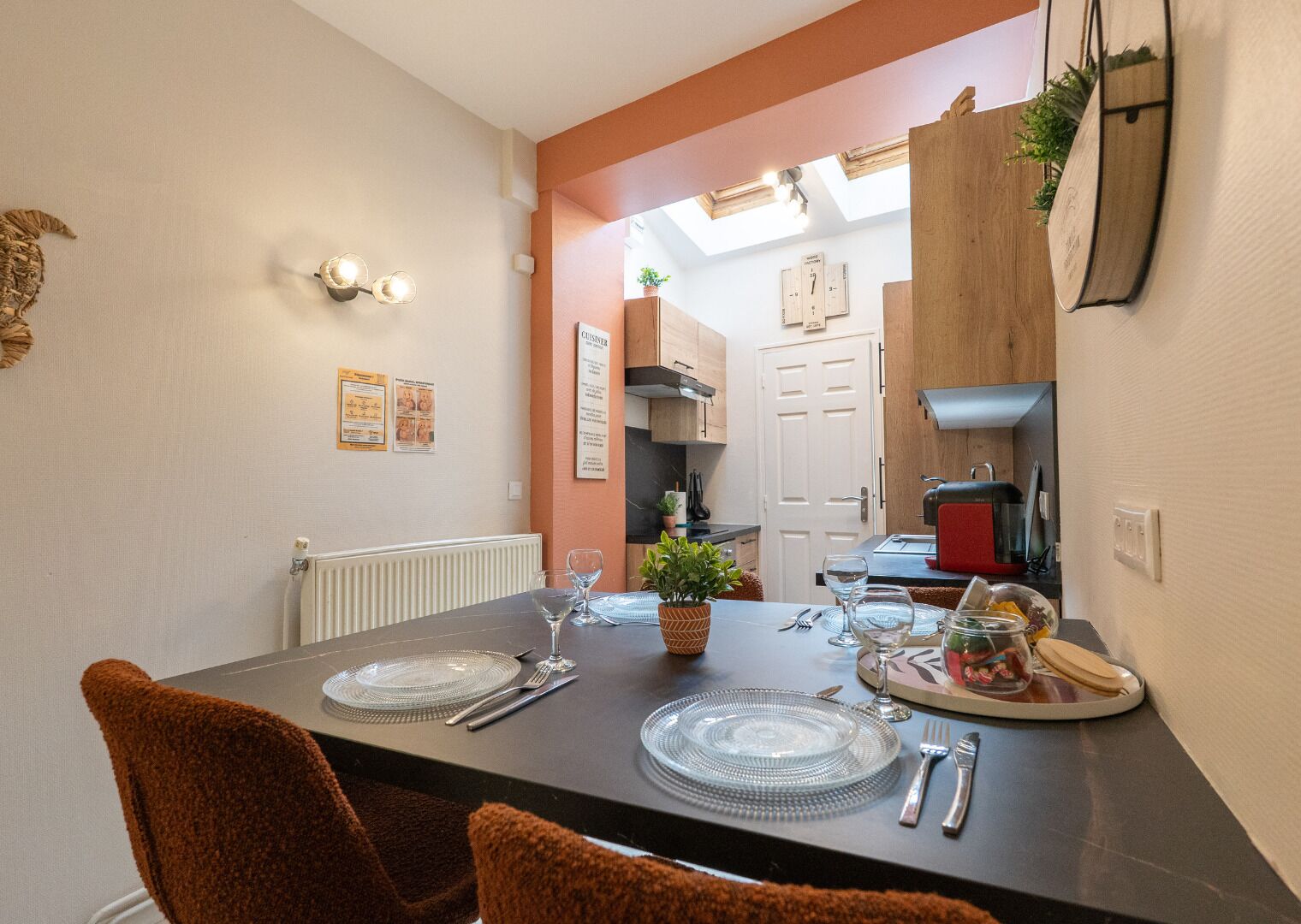 A dining area with a table set for two, a clock on the wall, and a view into the kitchen.