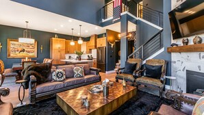 Ein geräumiges Wohnzimmer mit großem Couchtisch, Ledersofas und Kamin. Im Hintergrund ist eine Küche erkennbar, und eine Treppe führt auf eine obere Ebene.