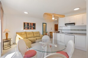 Un salon moderne avec une table à manger au plateau en verre, des chaises blanches, un canapé beige et un coin cuisine doté de meubles blancs.