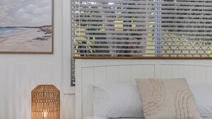 A bedroom with a bed, a bedside lamp, a window with blinds, and a painting on the wall. - Dreamtime Dune Studio (Fingal Head)