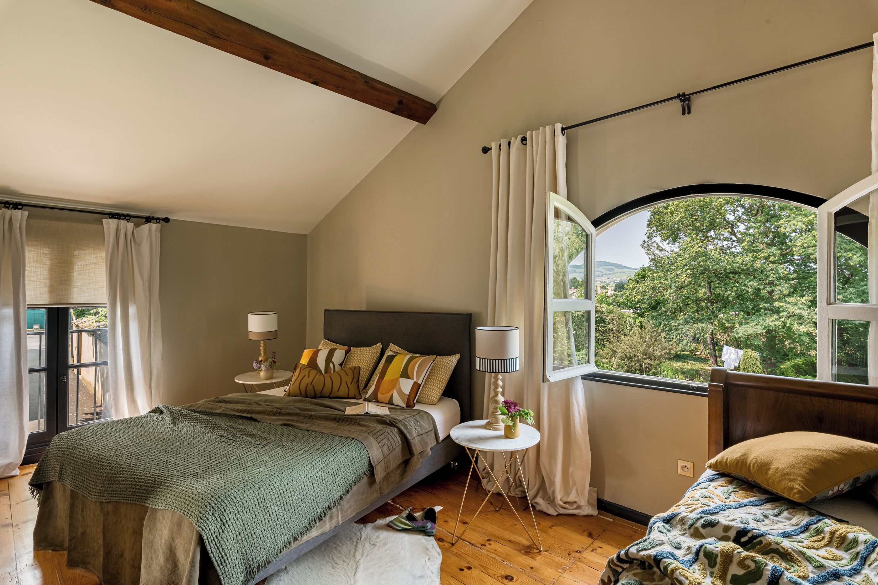A bedroom with a large bed, a smaller bed, a window with a view of greenery, and a small round table with a lamp.
