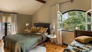 Ein Schlafzimmer mit einem großen Bett, einem Einzelbett, einem Fenster mit Blick auf Grünflächen und einem kleinen runden Tisch mit Lampe.