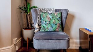 Een hoekkamer met een grijs gestoffeerde stoel, een kussen met een patroon, een potplant en een spiegel aan de muur.