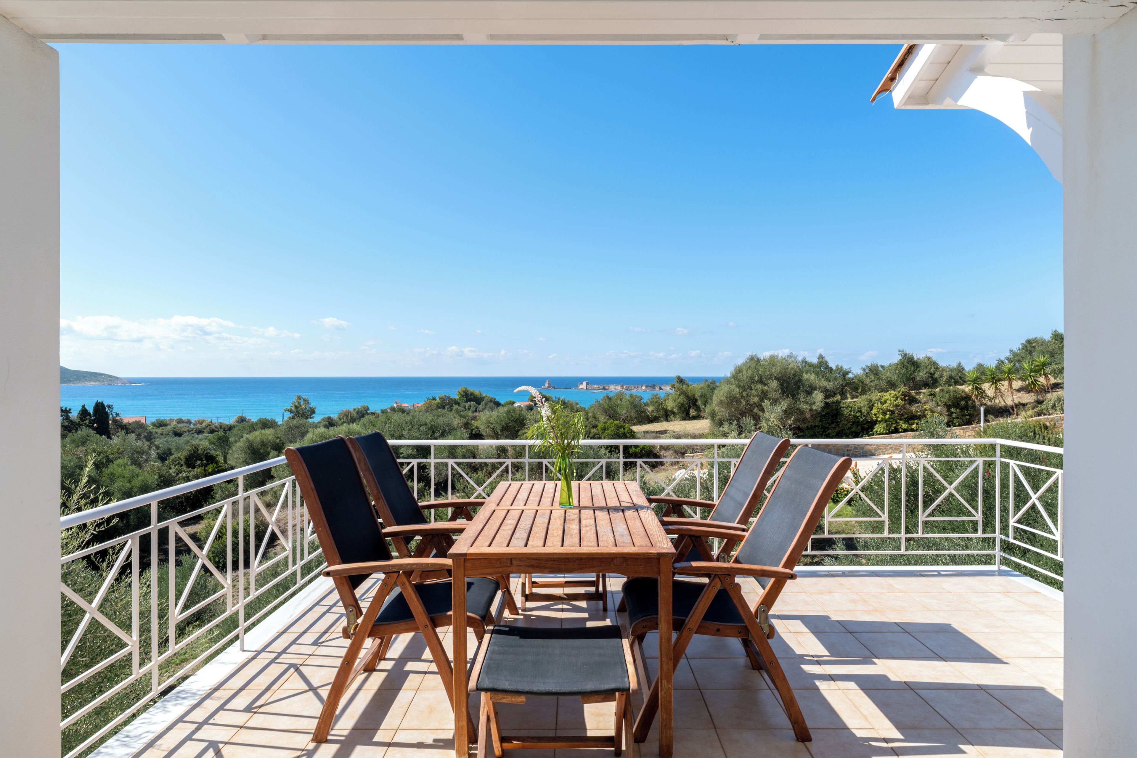 A wooden table and chairs on a balcony with a white railing, overlooking a scenic view of the sea and greenery.