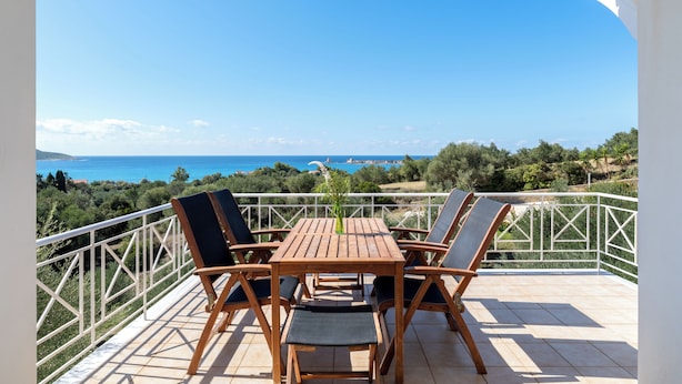 Una mesa y sillas de madera en un balcón con barandal blanco, con vista a un paisaje marino y de vegetación.
