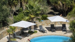 Zona de piscina con sillas de descanso y sombrillas, rodeada de muros de piedra y palmeras.