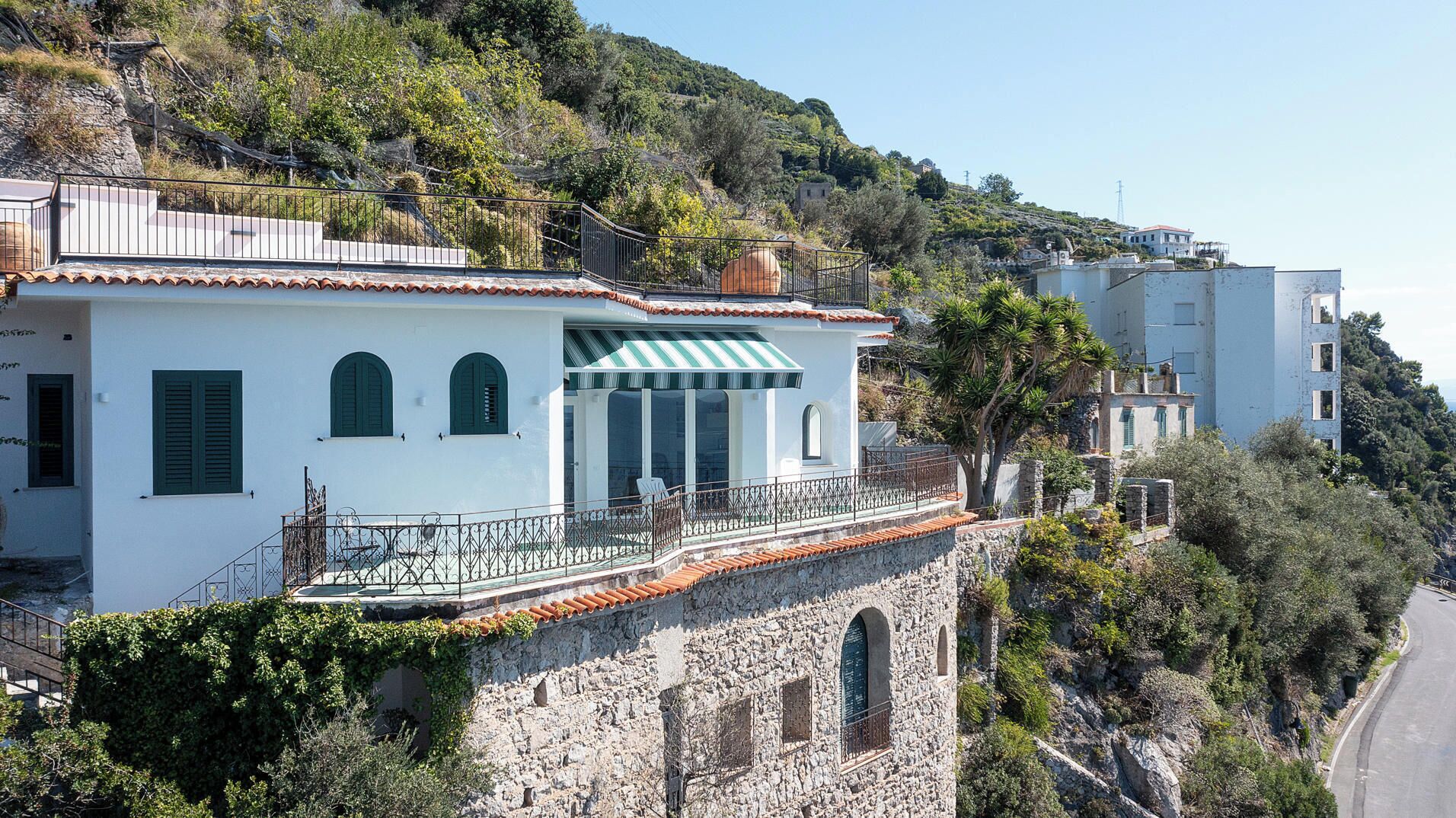 A white building with green shutters, a stone wall, and a balcony with a metal railing.