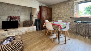 Ein gemütliches Zimmer mit Steinwand, einem Holztisch mit Stühlen, einem Fernseher und einem Fenster mit Blick auf Grünflächen.
