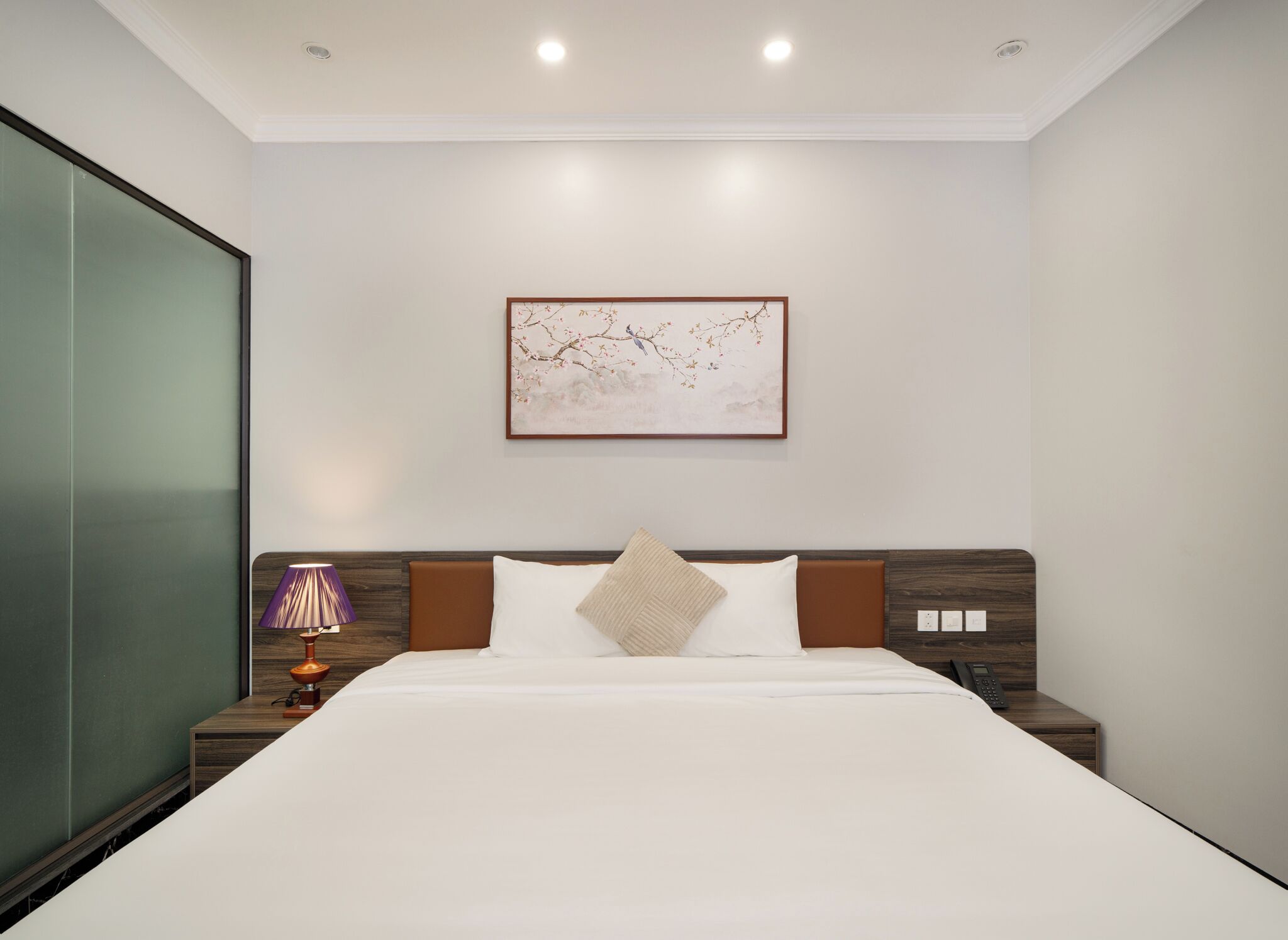 Une chambre d’hôtel moderne avec un grand lit, une table de chevet avec une lampe, un tableau accroché au mur et une tête de lit en bois.