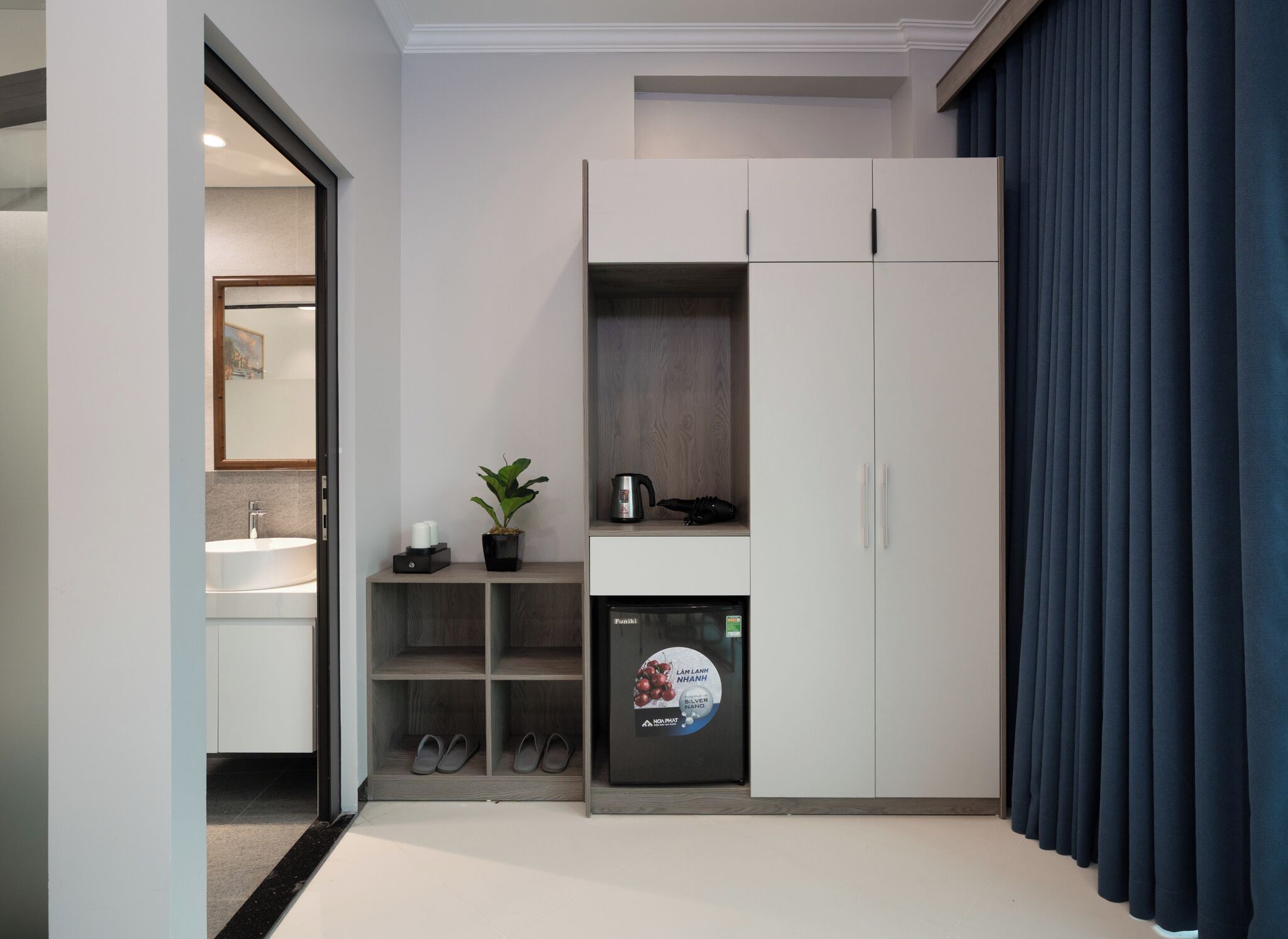 A modern room with a built-in fridge, a small shelf with a plant, a coffee maker, and a pair of slippers.