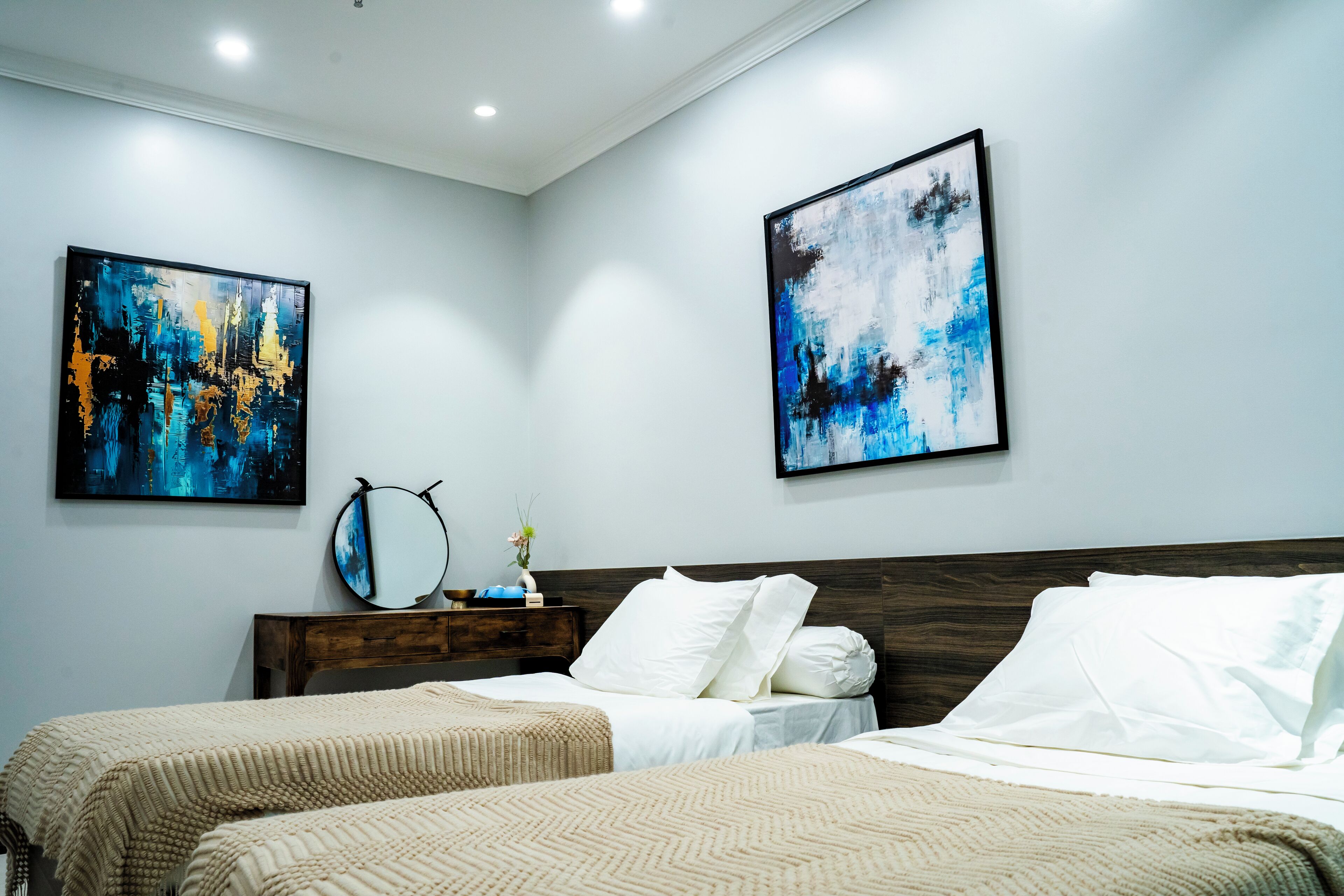 A hotel room with two beds, a wooden headboard, a nightstand with a mirror, and abstract art on the walls.