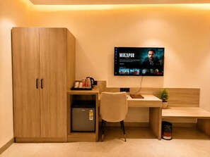 A hotel room with a wooden wardrobe, a desk with a chair, a small fridge, a television mounted on the wall, and a potted plant. - Prince Diamond Boutique Hotel (Varanasi)