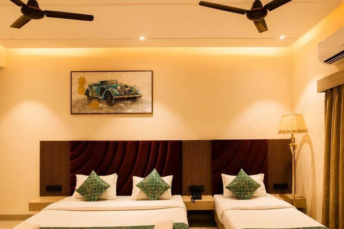 A hotel room with two beds, a ceiling fan, an air conditioning unit, a painting of a car, and a lamp.