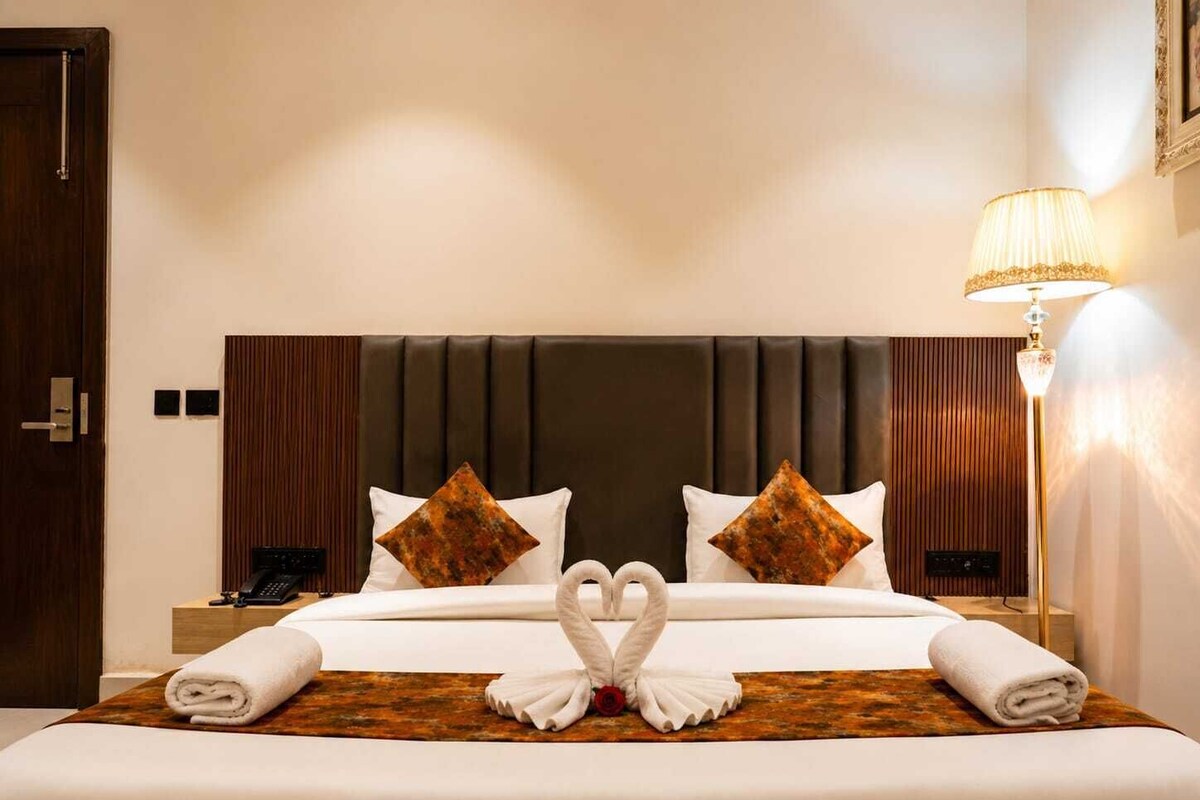 A hotel room with a large bed, two pillows, a bedside table with a lamp, and a door.