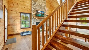 A wooden interior with a stone fireplace, a staircase, and a window overlooking greenery. - Valley View Cabin at Trickle Creek (Logan)