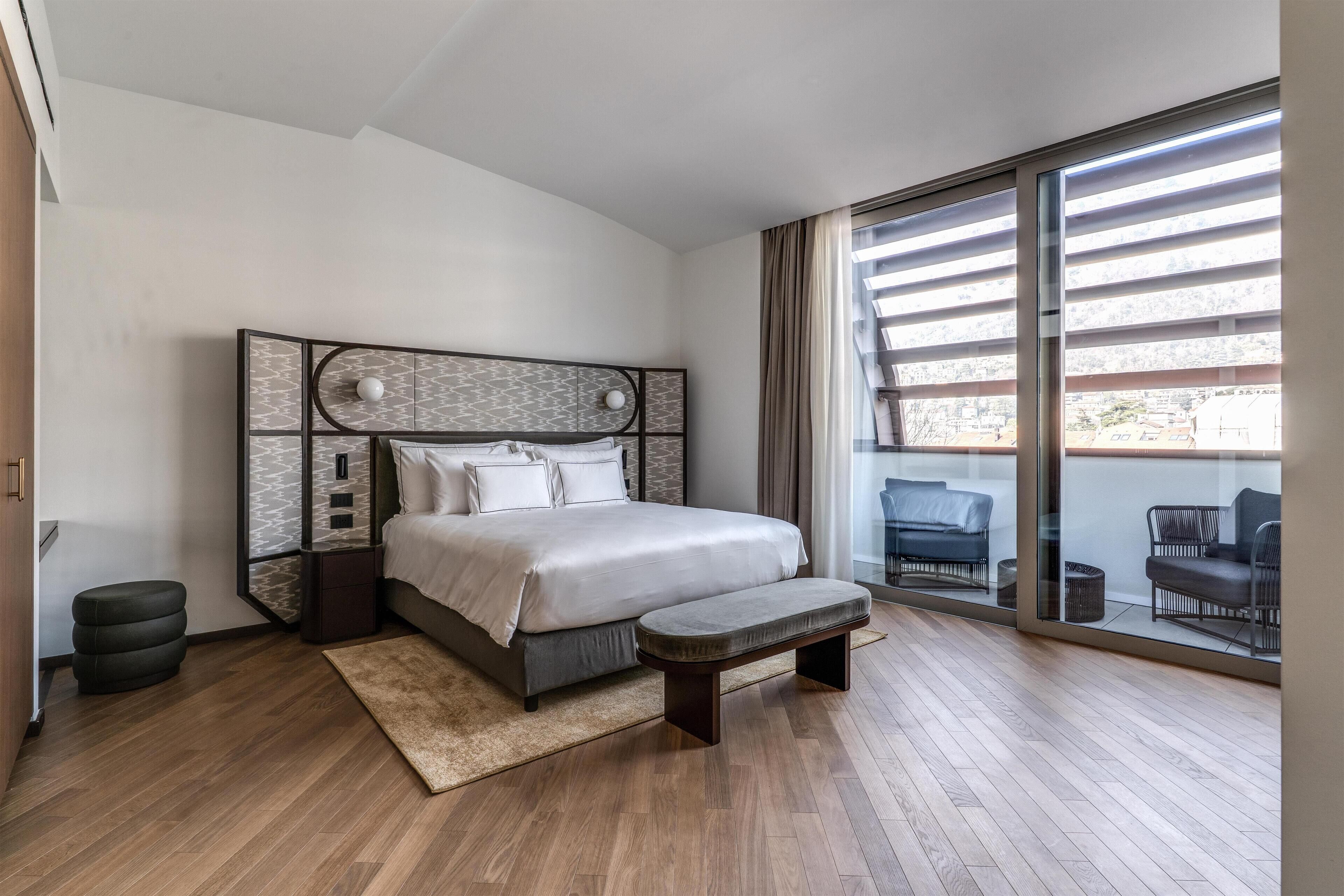 Un dormitorio moderno con una cama grande, un banco, un pequeño reposapiés y vistas a muebles de exterior a través de una puerta corrediza de vidrio.