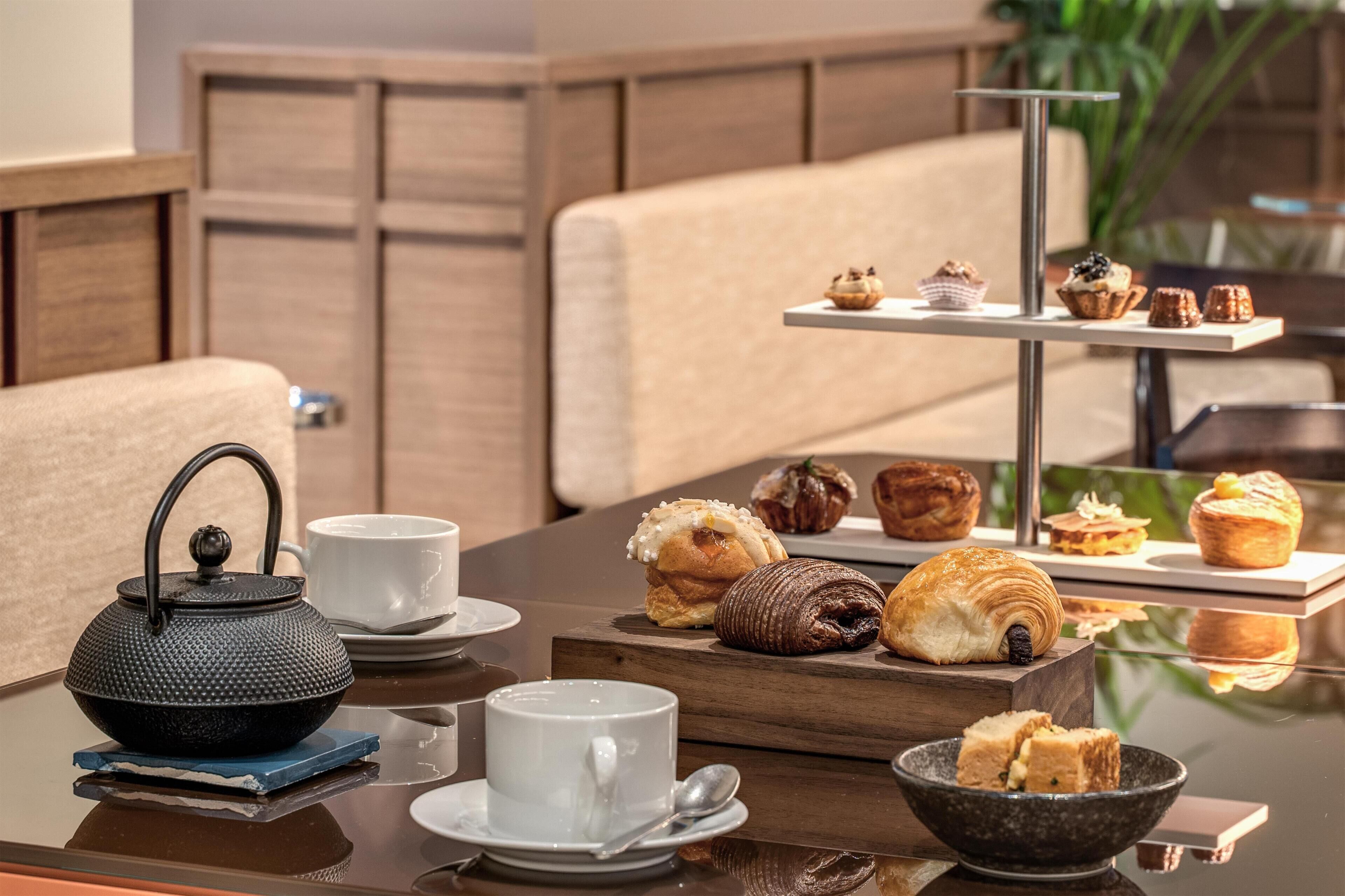 Una mesa con un calentador de té, tazas, pasteles y una bandeja de postres.