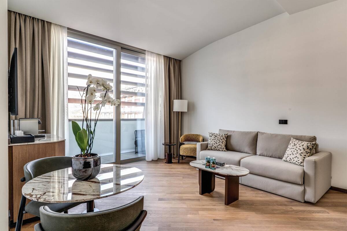 A modern living room with a sofa, a coffee table, a dining table with chairs, a potted plant, a lamp, and a TV.