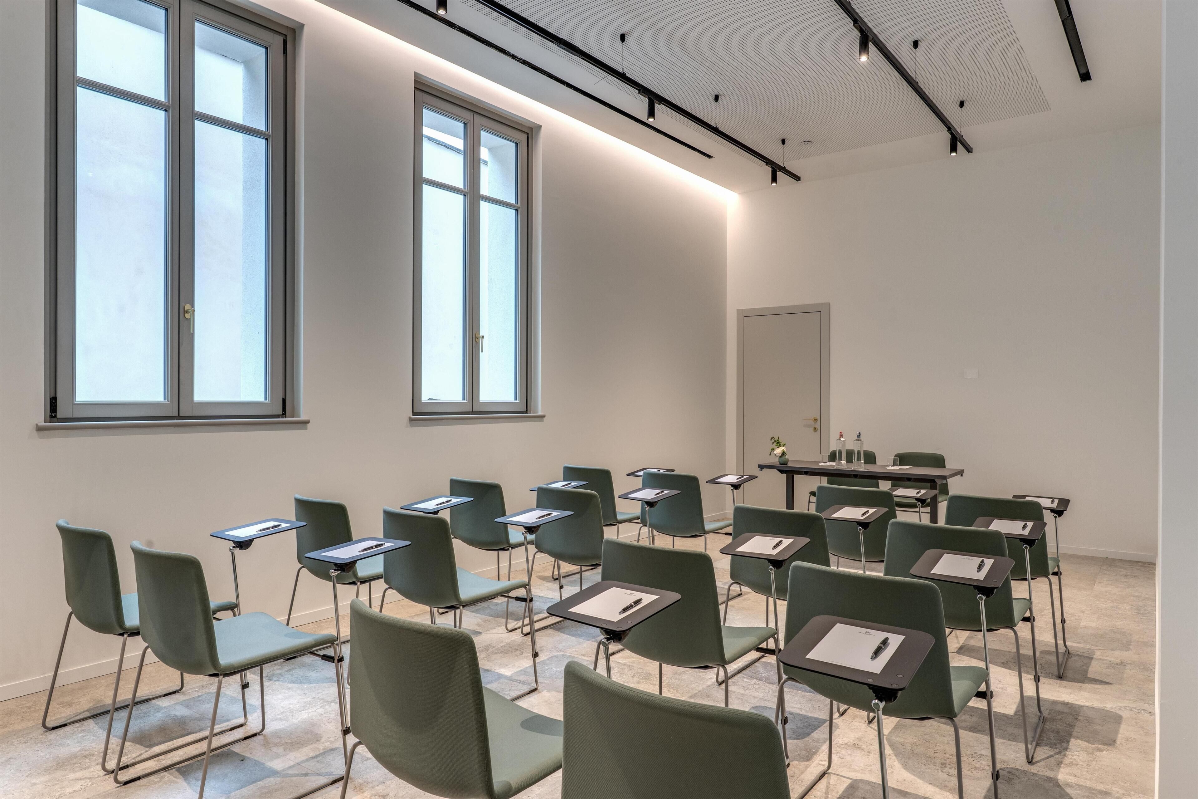 A classroom with green chairs and tables, large windows, and a white ceiling.