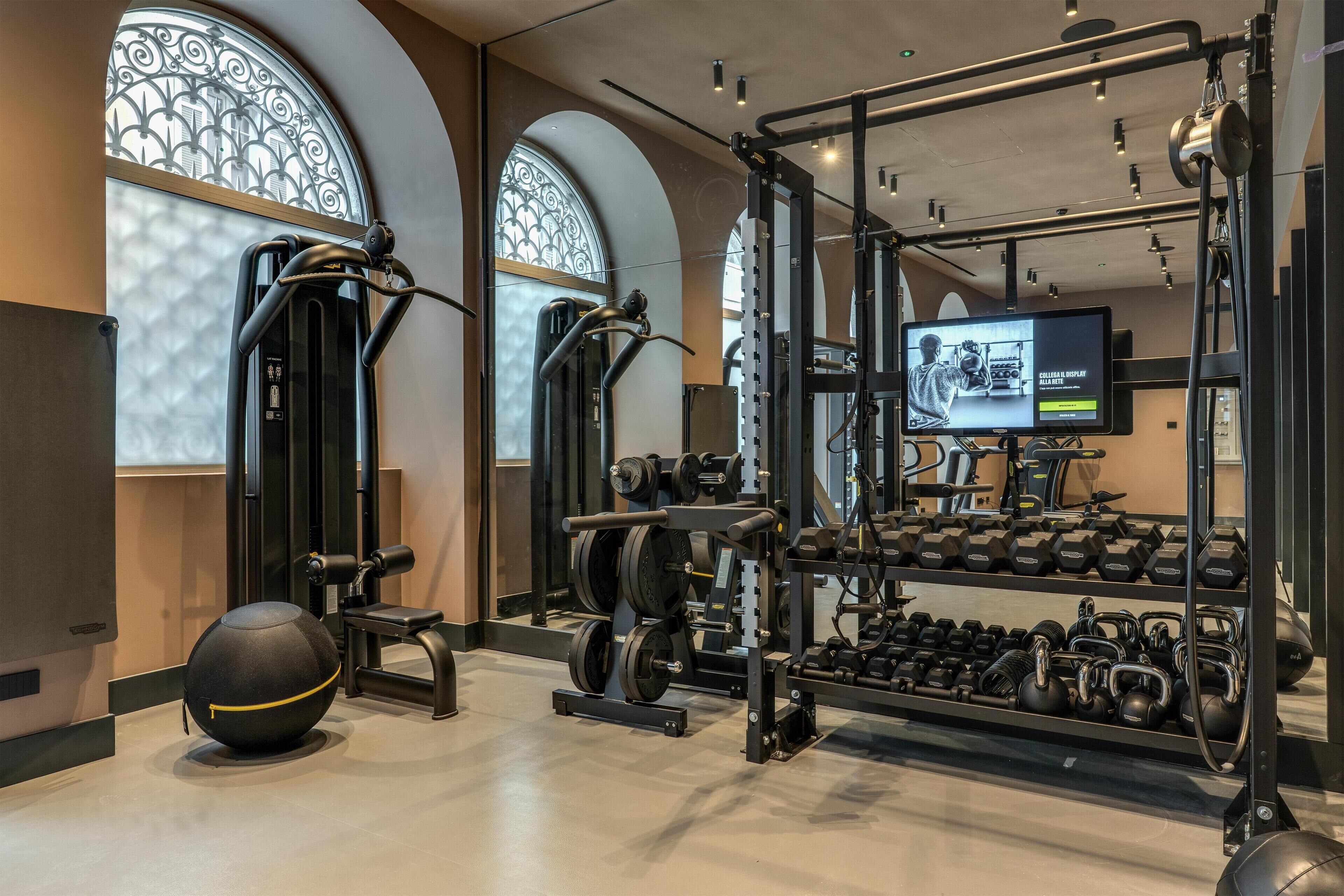Un gimnasio moderno con equipo de levantamiento de pesas, una pantalla de televisión y un ventanal con detalles de hierro forjado.