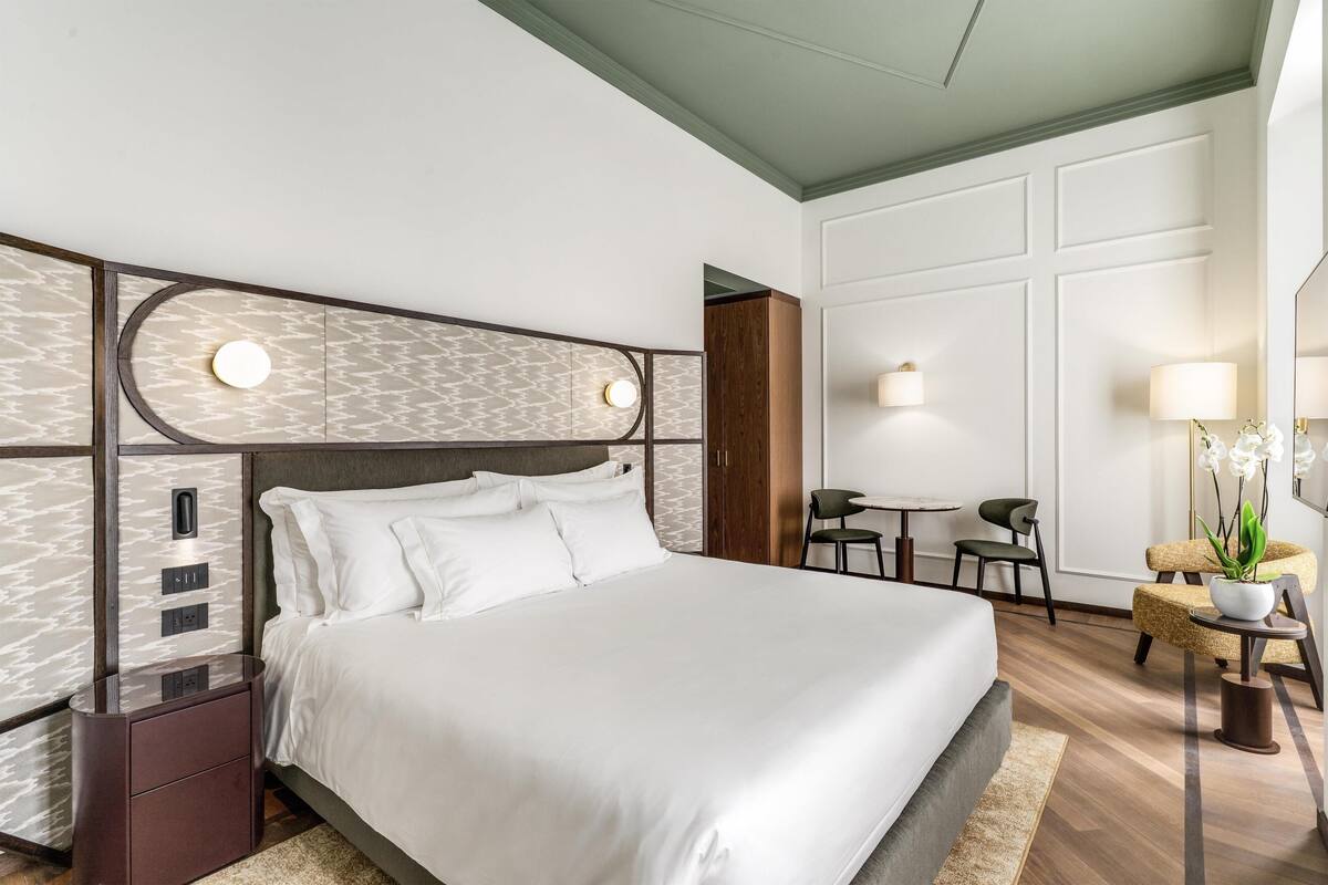 A modern hotel room with a large bed, a bedside table, a desk, and a chair.