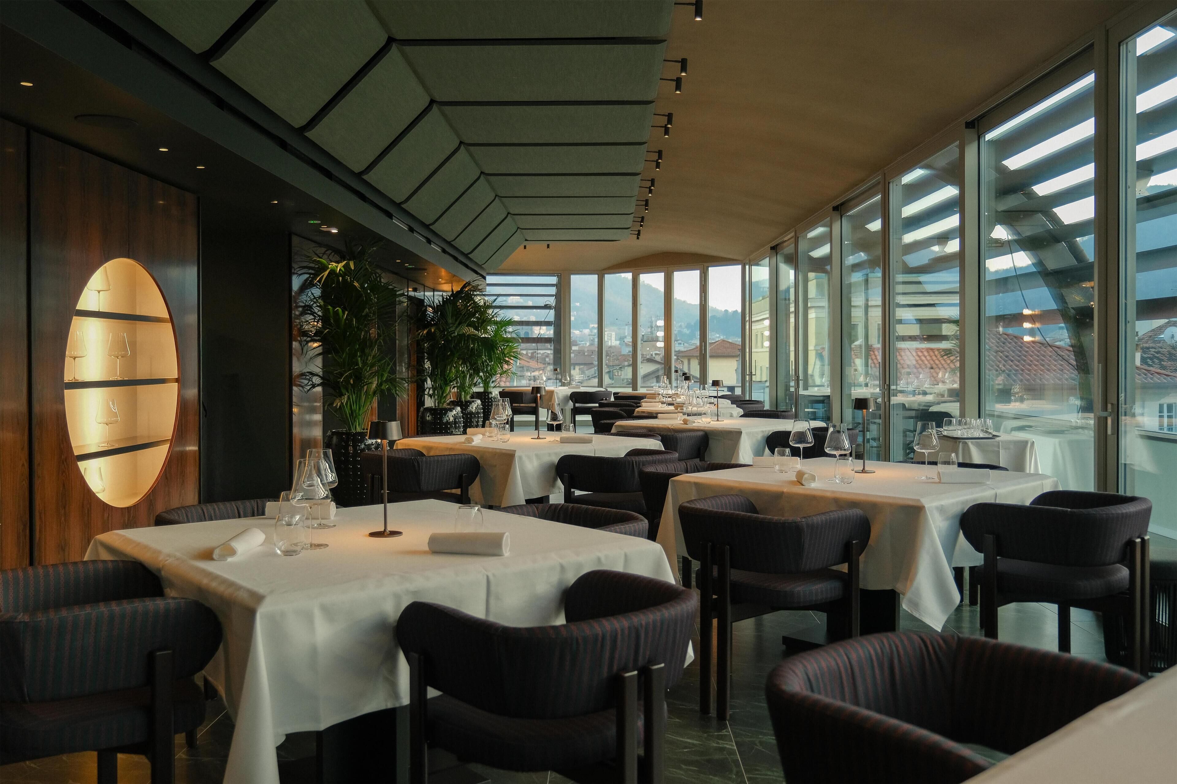 Een restaurant met meerdere tafels en stoelen, grote ramen en uitzicht op de stad.