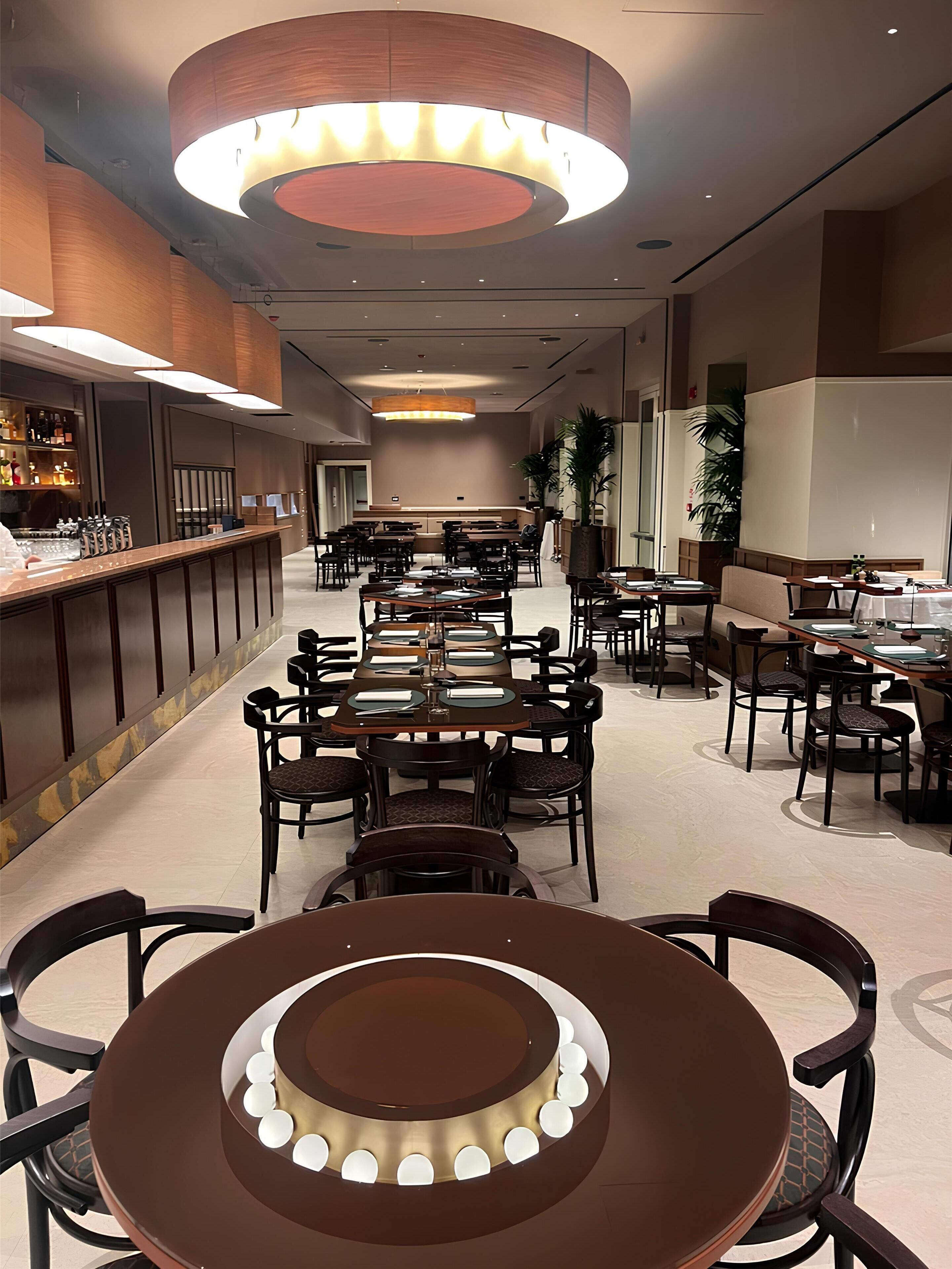 Un amplio vestíbulo de hotel con varias mesas y sillas para comer, una zona de bar y una gran lámpara circular en el techo.