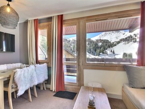 Ein Zimmer mit einem Holztisch, einer Couch, einem Sessel mit weißer, flauschiger Decke, einem Fernseher und Blick auf eine verschneite Berglandschaft.