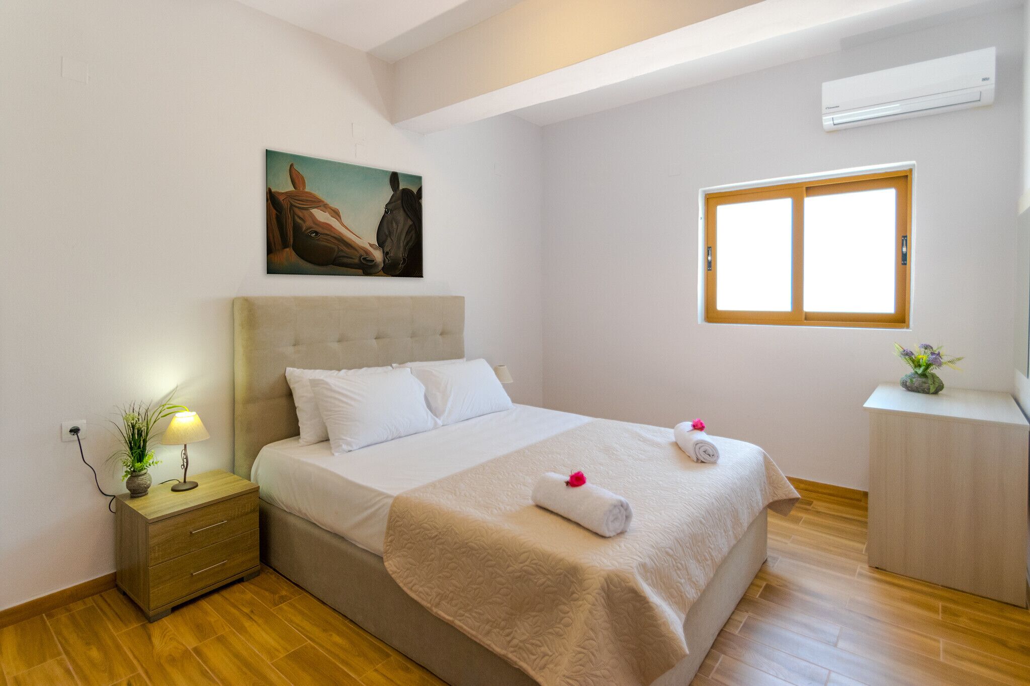Uma cama bem arrumada com cabeceira, mesa de cabeceira com abajur e planta, janela com persianas e um quadro na parede.