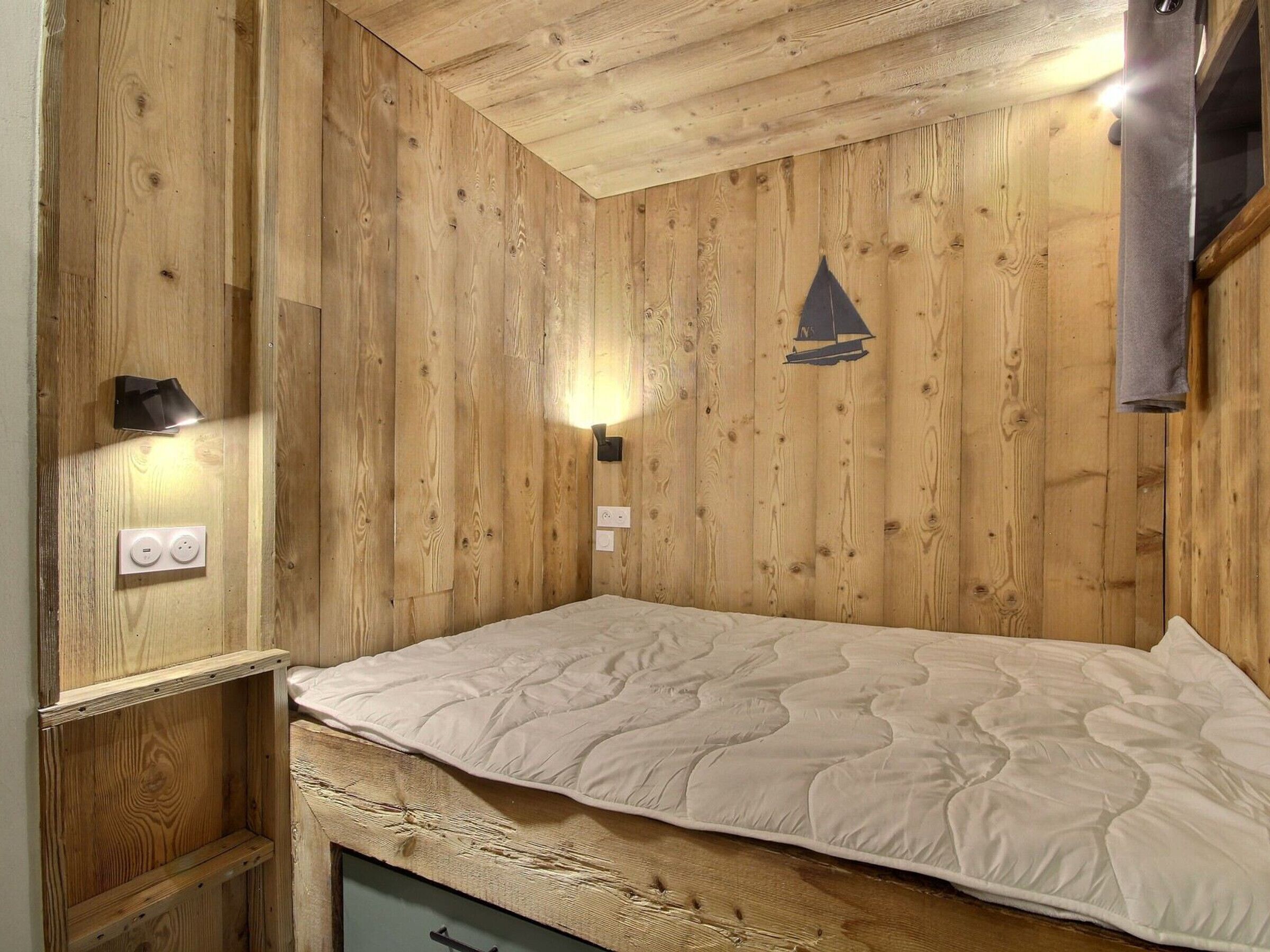 A wooden-paneled room with a bed, a wall-mounted light, and a small shelf.