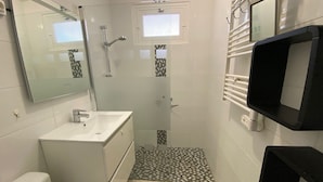 Une salle de bain avec un lavabo blanc, un miroir et des toilettes. Il y a un espace douche avec un sol carrelé et une fenêtre située au-dessus de la douche.